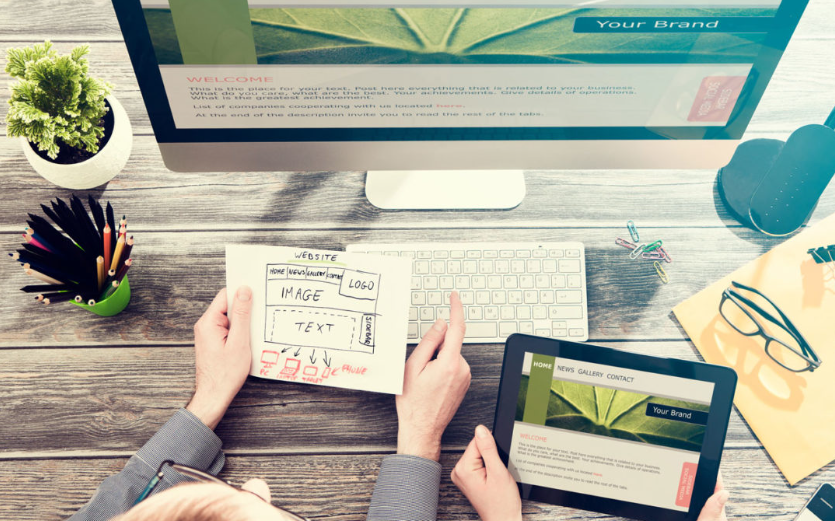 Two people collaborate at a wooden desk, immersed in web development. A computer monitor displays a site, with one person holding a wireframe sketch and the other using a tablet to view a webpage. Nearby are glasses, office supplies, and a plant; the scene captures the essence of Melbourne's vibrant web design agency culture. | Ven Agency